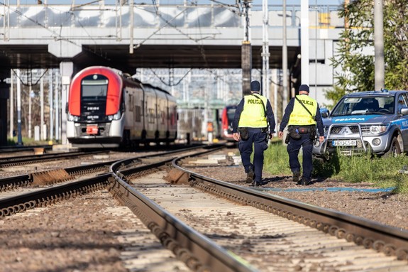 Dwaj umundurowani policjanci widziani z tyłu patrolują rejon torowiska kolejowego. Na drugim planie szynobus po lewej stronie i radiowóz po prawej.