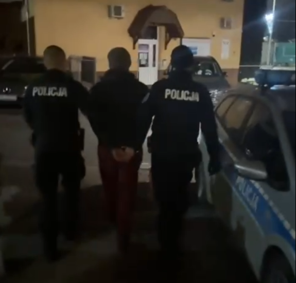 Sceneria nocna. Dwaj policjanci prowadzą zatrzymanego idąc z nim do radiowozu stojącego przy budynku komendy policji.