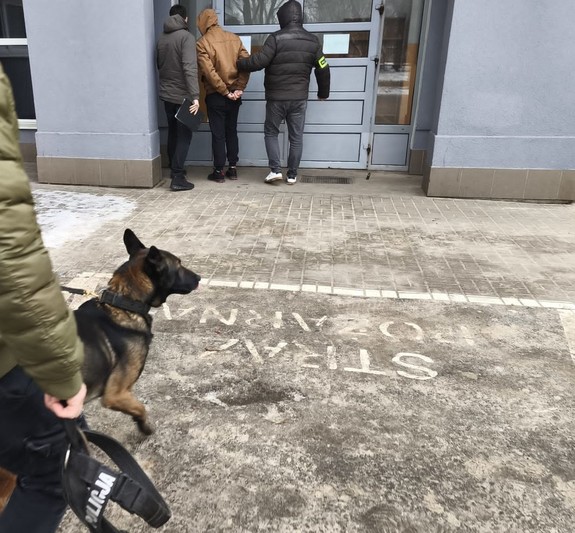 Dwaj nieumundurowani policjanci stoją z zatrzymanym przy drzwiach budynku. Za nimi policjant z psem.